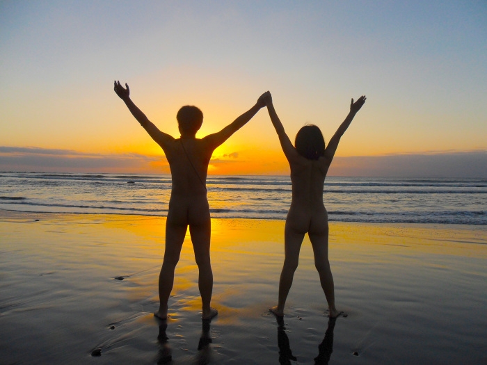 Couple at a beach, raising arms to the sunrise. Sunday Nudist.