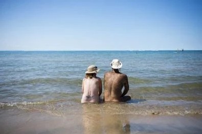 Two older, nude people sitting at a beach