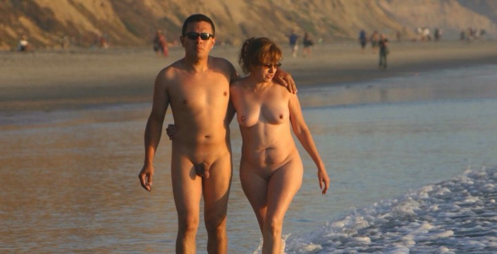 2 nude people walking in the water along a beach